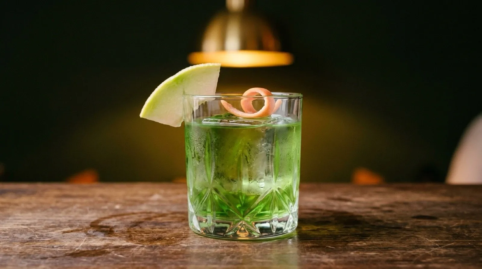 STILLROOM Madrid Jerónimos signature Green Negroni: translucent green cocktail in a carved rocks glass with a large ice cube, a slice of galia melon and a pink grapefruit twist