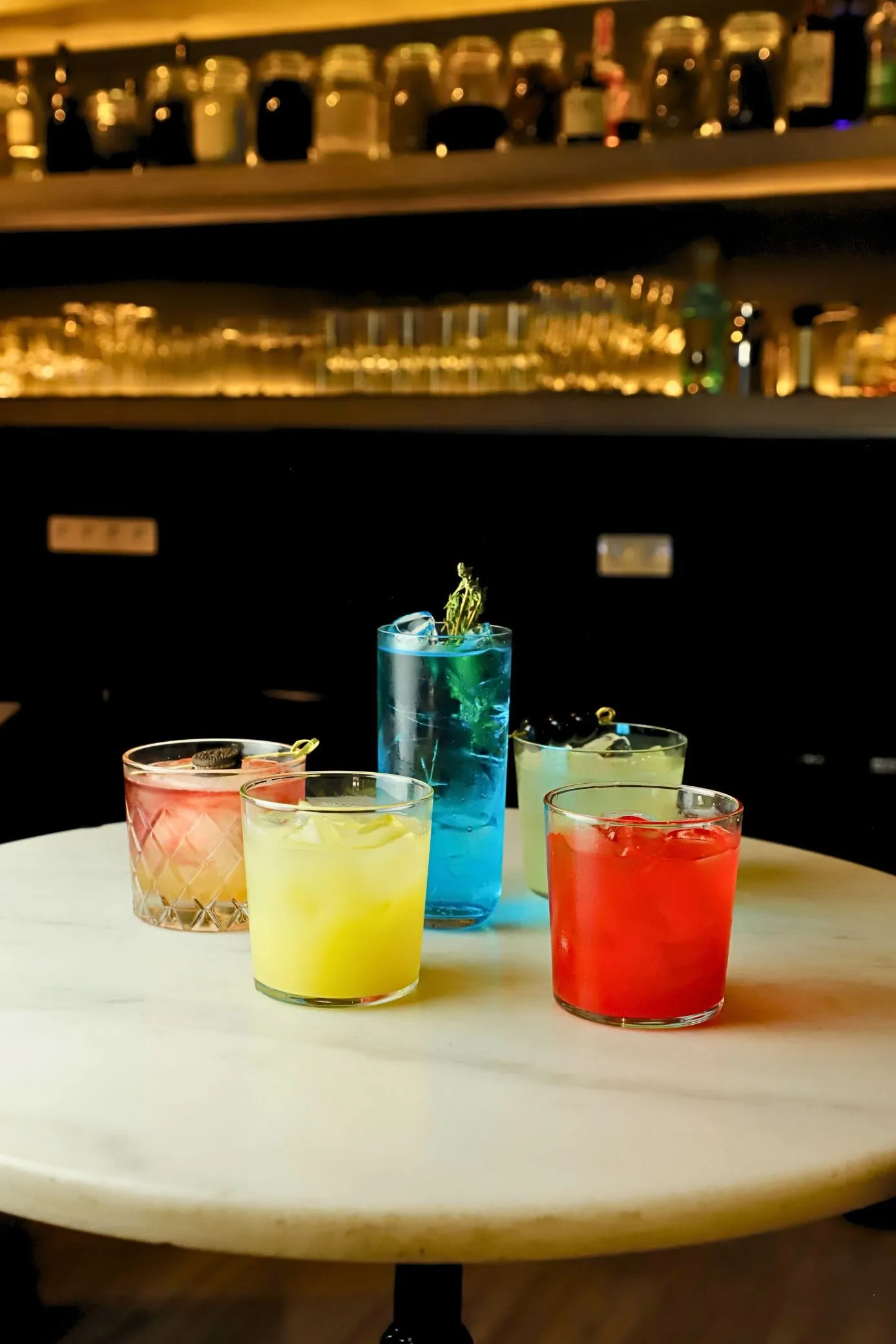 Cinco cócteles de colores sobre la mesa de mármol de Stillroom, con la barra de tarros iluminada al fondo