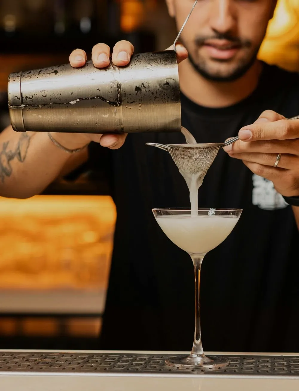 Un bartender colando un cóctel blanco en copa coupé sobre la barra